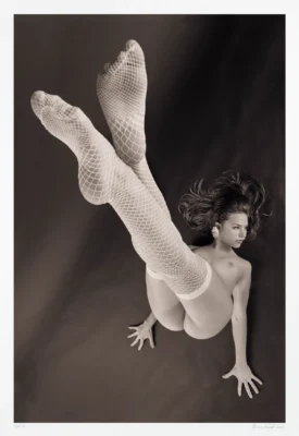 In this black and white photograph, a nude woman is depicted balancing, inverted and vertical, as seen from above. Fishnet stockings evoke classic pinup