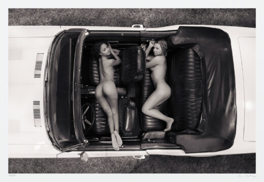 This black and white art photograph incorporates notions of freedom, Americana, and sensuality. A vintage fastback convertible, holds two sensually-posed women. The vantage is from directly above the middle of the vehicle. showing the nude figures between its two doors, dash, and drop-top.