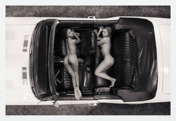 This black and white art photograph incorporates notions of freedom, Americana, and sensuality. A vintage fastback convertible, holds two sensually-posed women. The vantage is from directly above the middle of the vehicle. showing the nude figures between its two doors, dash, and drop-top.