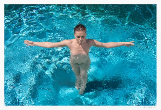 In this colorful art photograph, a nude woman stands in a swimming pool. She extends both arms symmetrically, forming a an unmistakable cross shape.